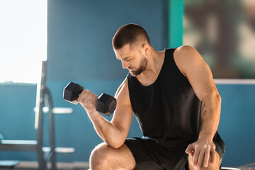 A man is seated on a bench in gym, gripping a pair of dumbbells in his hands. He appears focused and concentrated on his workout routine, showcasing determination and commitment to fitness.