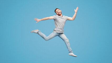 Funny millennial guy jumping in air and fooling on blue background, full length, empty space