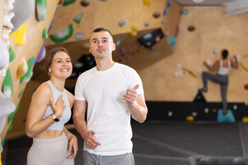 Interested young girl and man standing near bouldering wall in climbing gym, engaged in conversation. Coach instructing beginner female climber about specific moves or technique - obrazy, fototapety, plakaty