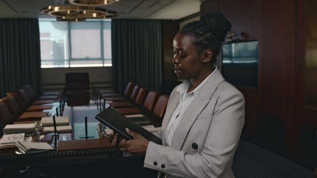 Push in portrait of positive female African American lawyer in grey suit posing with black leather folder in luxurious conference hall at law firm and looking at camera