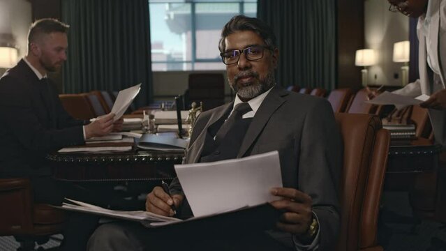 Medium portrait of senior Biracial law firm founder in suit and glasses posing in leather armchair with documents in conference hall and looking at camera, diverse attorneys working in background