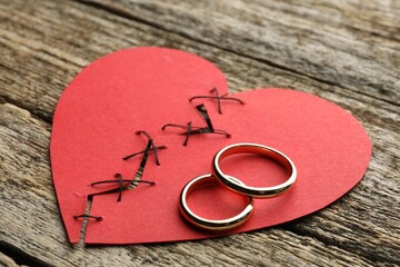 Broken heart. Torn red paper heart sewed with thread and wedding rings on wooden table, closeup