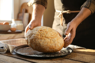 Man holding loaf of fresh bread at wooden table indoors, closeup