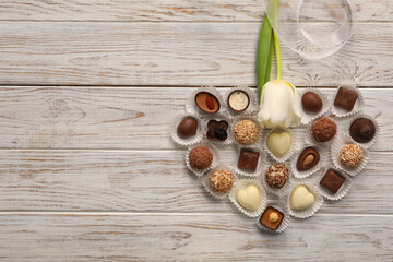 Heart made with delicious chocolate candies and beautiful tulip on white wooden table, flat lay. Space for text