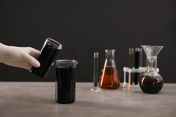 Woman pouring black crude oil into beaker at grey table against dark background, closeup