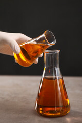 Woman pouring yellow crude oil into flask at grey table against dark background, closeup