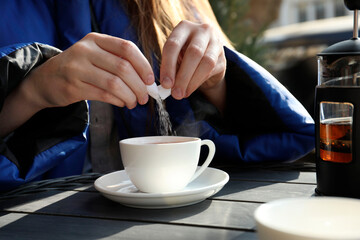Woman adding sugar into cup of tea at black wooden table in outdoor cafe, closeup