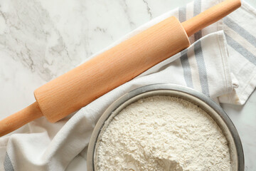 Flour in bowl, rolling pin and napkin on white marble table, top view