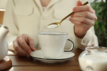 Woman adding sugar into aromatic tea at wooden table indoors, closeup