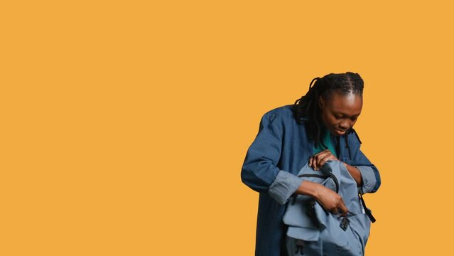 Joyous african american woman rummaging through backpack, looking for school notebooks, isolated over studio background. Merry teenager taking out university supplies out of rucksack, camera B