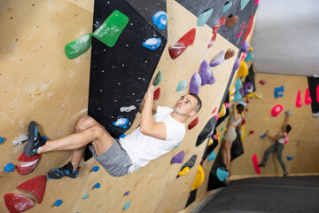 European man is mastering climbing on training wall in gym, side view. He holds on tightly to...