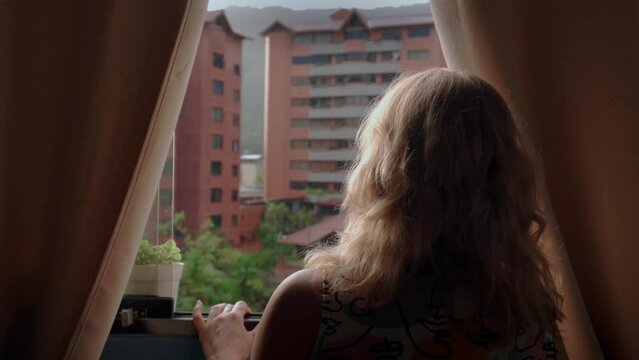 Mujer rubia con ropa de casa mirando por la ventana que est&aacute; lloviendo