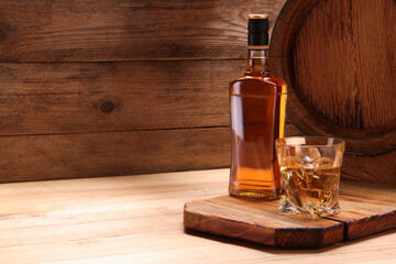 Whiskey with ice cubes in glass, bottle and barrel on wooden table, space for text