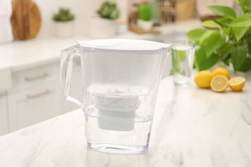Water filter jug, glass and lemons on white marble table in kitchen