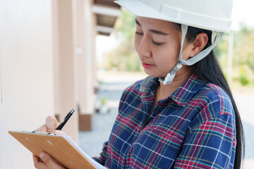 Close-up of Asian female inspector in safety helmet, clipboard in hand, writing notes, eyes...