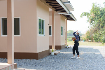 Asian female in safety helmet, gazing upwards, standing outside, clipboard in hand, inspecting newly constructed house. Inspectors inspect the completed house.