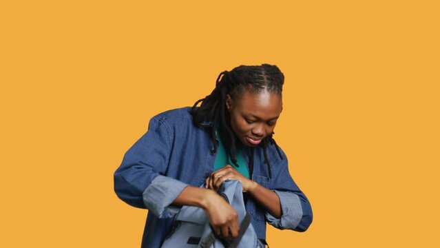 Joyous african american woman rummaging through backpack, looking for school notebooks, isolated over studio background. Merry teenager taking out university supplies out of rucksack, camera A