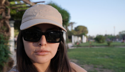 Portrait of a young confident woman wearing sunglasses and a cap, in the park at sunset 

