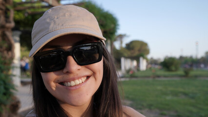 Portrait of a young woman wearing sunglasses and a cap, smiling at camera, in the park at sunset