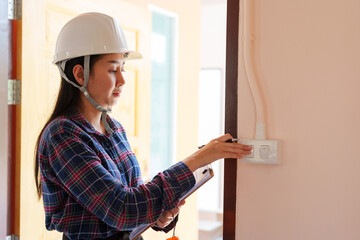 Asian female in plaid shirt, white safety helmet, inspecting electric switch, with clipboard,...