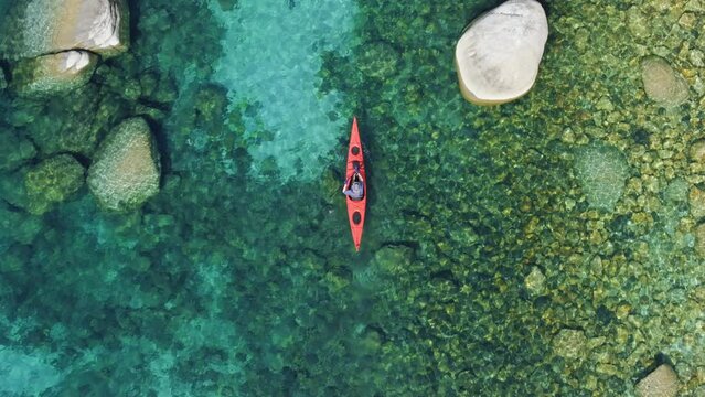 A birds eye view of a person in a red kayak on the water, surrounded by green shrubs on the grassy shoreline. Underwater plants peek through the clear water