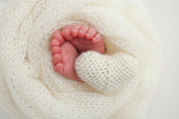 The tiny foot of a newborn baby. Soft feet of a new born in a white wool blanket. Close up of toes, heels and feet of a newborn. Knitted white heart in the legs of a baby. Macro photography