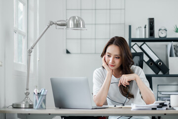 Young businesswoman appears reflective while seated at her bright and orderly home office, with a laptop and documents on her desk.