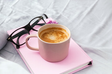 Composition with pink mug with coffee, pink notepad and glasses on the bed.