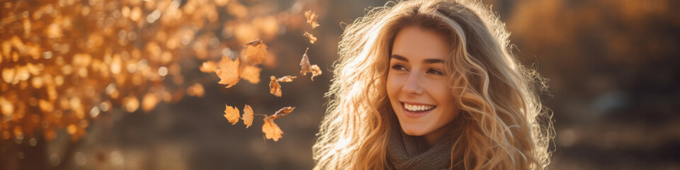 Fototapeta premium A woman with long blonde hair is smiling in front of a tree with leaves falling
