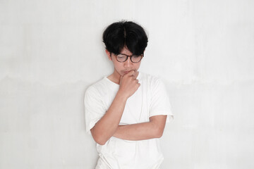 asian man in casual clothes and glasses thinking and doubting, holding his chin with hand on white...