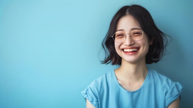 Cheerful young Asian woman with short black hair, wearing round glasses and a light blue t-shirt. Bright, genuine smile radiates happiness and friendly approachability. 