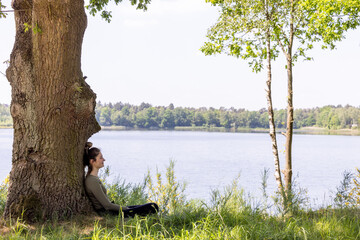 An image of quiet repose, showing a woman seated under the sturdy trunk of a tree. The lake in the...