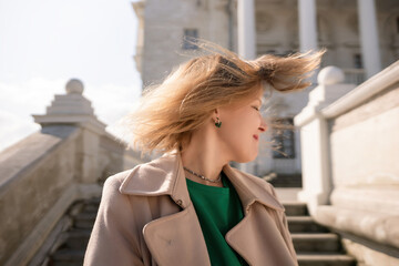 A woman with long hair is standing on a set of stairs. She is wearing a green shirt and a tan coat.