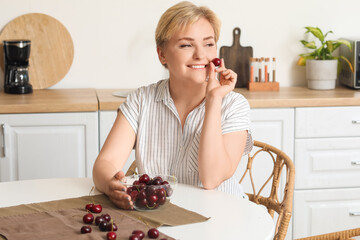Beautiful young happy woman with bowl of ripe cherries sitting at table in kitchen