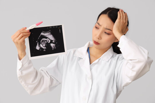 Upset female Asian doctor with pregnancy test and sonogram image on light background