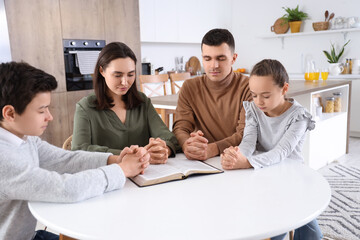 Family praying with Holy Bible on table in kitchen