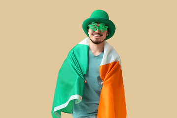 Happy young man in leprechaun's hat and party glasses with flag of Ireland on beige background. St....