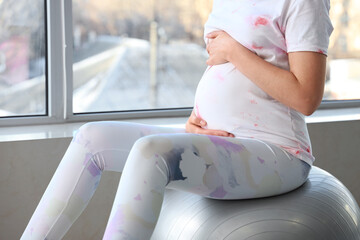 Young pregnant woman sitting on fitball near window at home