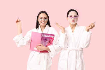 Young women in bathrobes with facial mask, under-eye patches, lip balms and magazine on pink background