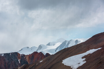 Awesome aerial view to sharp rocks on ridge of vivid colors and snow-white peak in low clouds. Scenic misty landscape with colorful sheer crags against snow-capped mountain top under low cloudy sky.