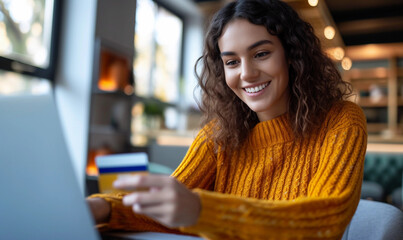 Pretty student girl surfing internet in home, sitting at table with open laptop and mug, holding plastic credit card, entering information via paying system while shopping online.