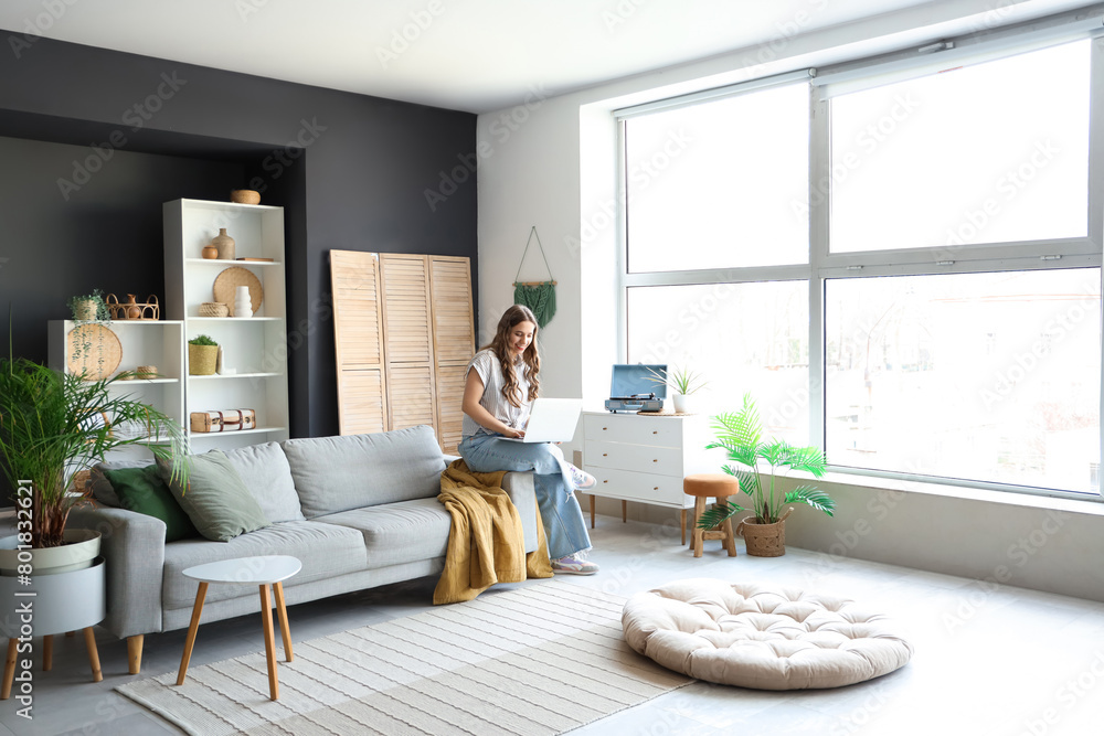 Wall mural Young woman using laptop on grey sofa at home