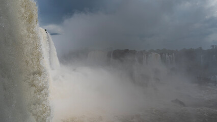 The powerful waterfall is shrouded in fog. Clouds in the blue sky. In the foreground, the flow of...