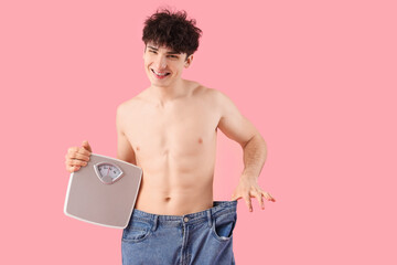 Handsome happy young sporty man in loose jeans with scales on pink background. Weight loss concept