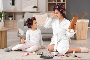 Little Asian girl and her mother with hair curlers doing makeup at home