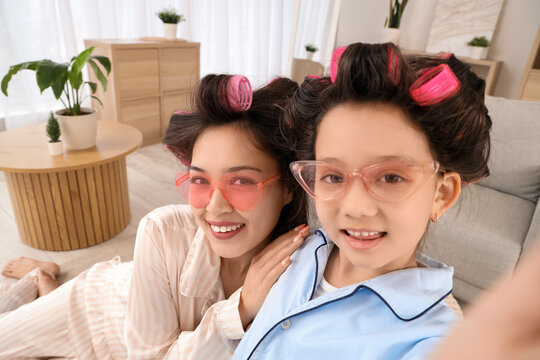 Little Asian girl and her mother in sunglasses with hair curlers taking selfie at home, closeup
