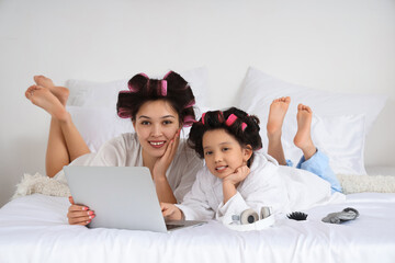 Cute little Asian girl and her mother with hair curlers using laptop in bedroom