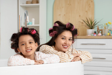 Little Asian girl and her mother with hair curlers in bathtub at home