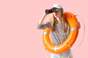 Young female lifeguard in cap with rescue ring looking in binocular on pink background