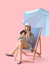 Young female lifeguard in cap sitting on deck chair with binocular near umbrella on pink background